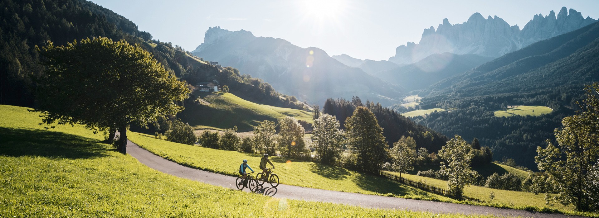 The picture shows two cyclists riding under the Dolomites or the Villnöss Geislern on a narrow road through a green meadow.