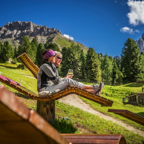 The picture shows a couple resting on deck chairs made of pure wood on an alpine pasture in the middle of the green meadow.