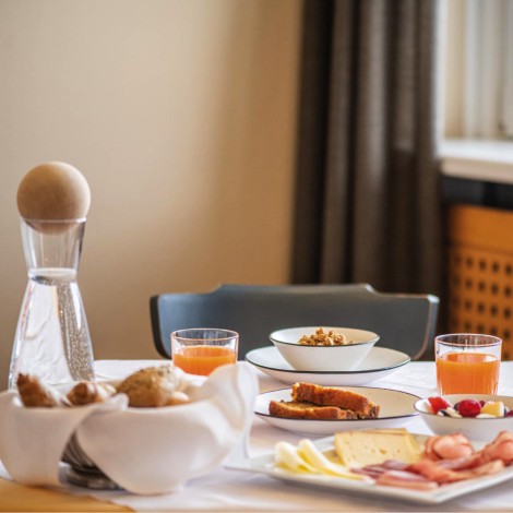 Here you see a richly laid breakfast table. There are many products on it: orange juice, fresh bread, sausage, cheese and a water carafe with a wooden stone-pine ball on top.