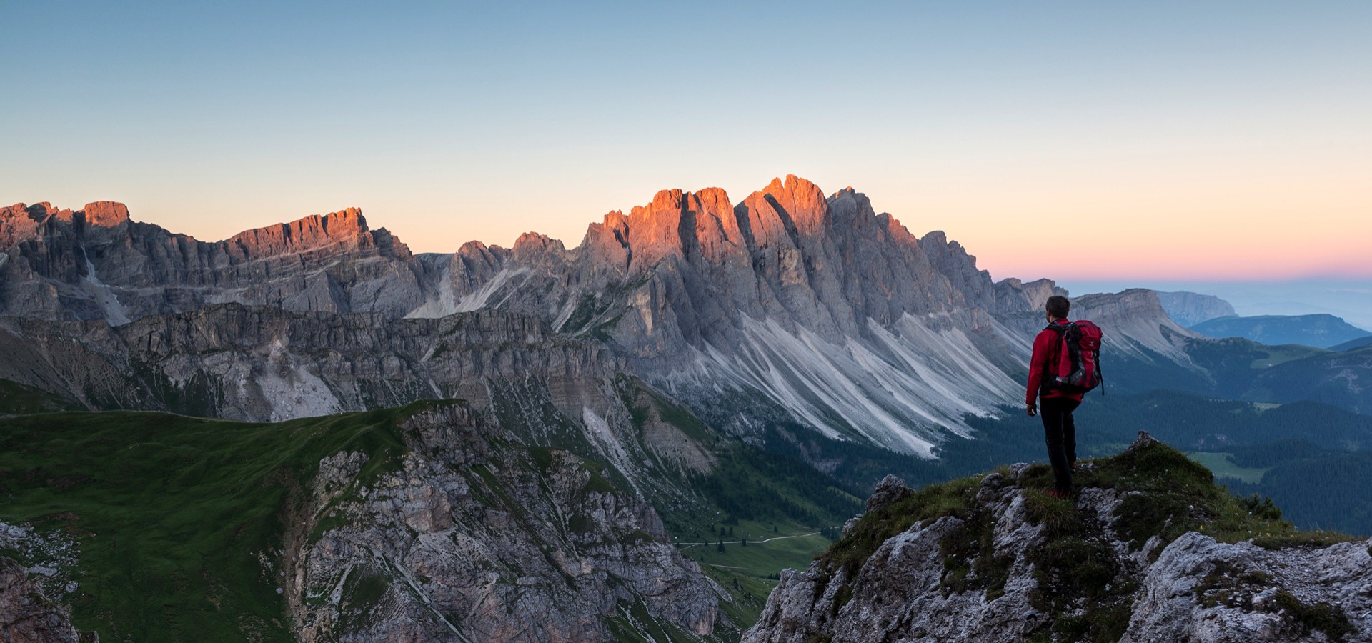 The picture shows a walking man resting at a vantage point and admiring the Dolomites in the sunset.