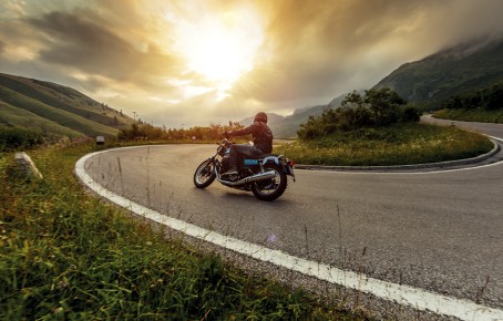 The picture shows a man riding a motorcycle along the pass road. The sun is breaking through the clouds.