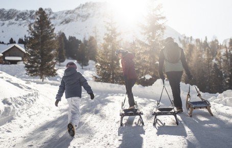 The picture shows a family on a hike through the South Tyrolean winter landscape, pulling a toboggan behind them.