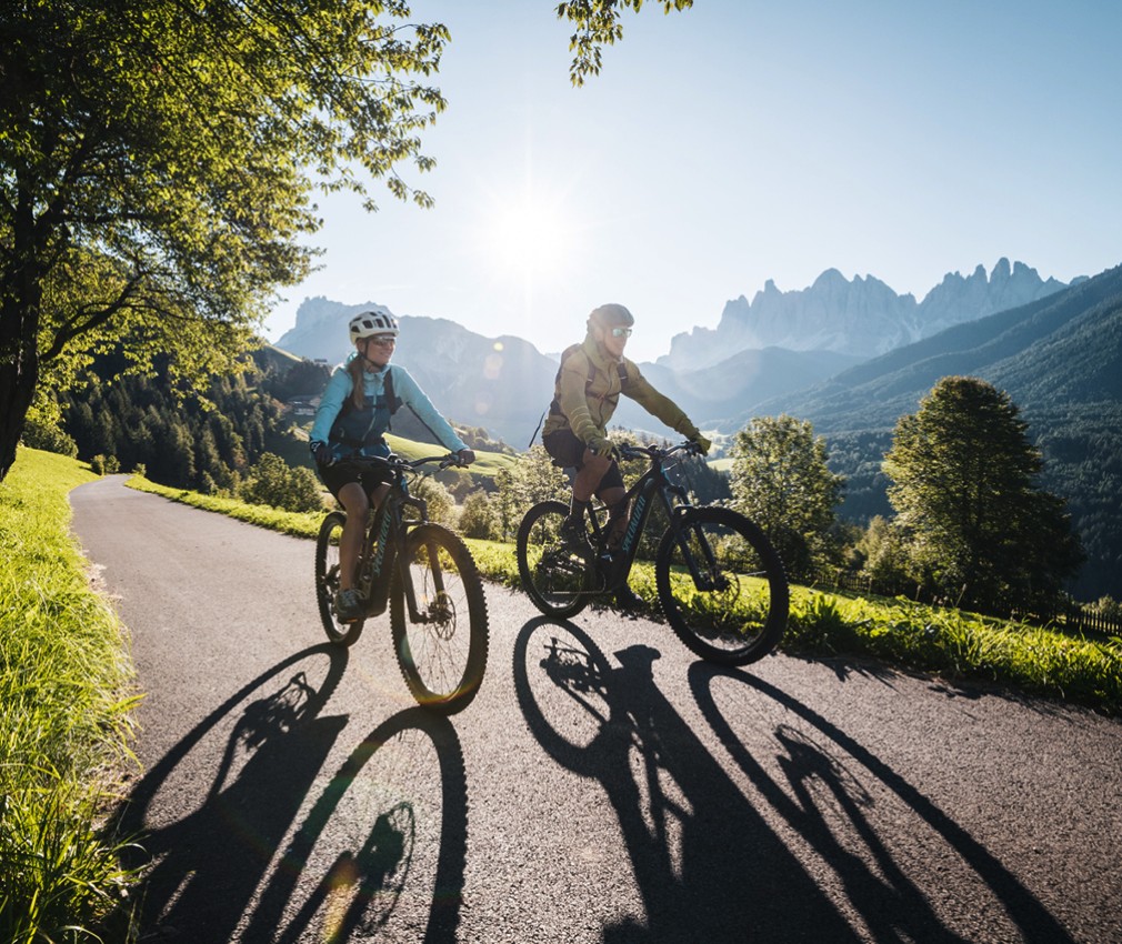 The picture shows two cyclists riding under the Dolomites or the Villnöss Geislern on a narrow road through a green meadow.