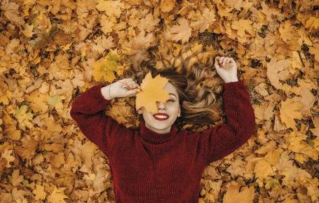 Das Bild zeigt eine Frau, die zwischen den Herbstblättern liegt und sich eines ins Gesicht hält. Sie lächelt.