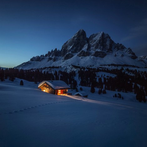 The picture shows a rustic wooden mountain hut at dusk under the Peitlerkofel. Warm light burns in the hut.